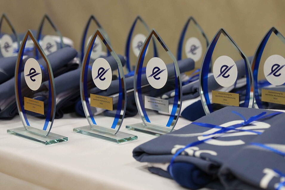 Troph&auml;en in Tropfenform mit blauen Akzenten stehen neben gestapelten T-Shirts auf einem Tisch.