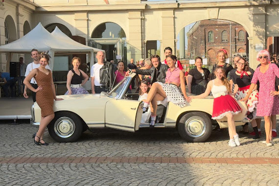 Gruppe von Menschen in Retro-Kleidung posiert um ein wei&szlig;es Oldtimer-Cabrio auf einem Platz.