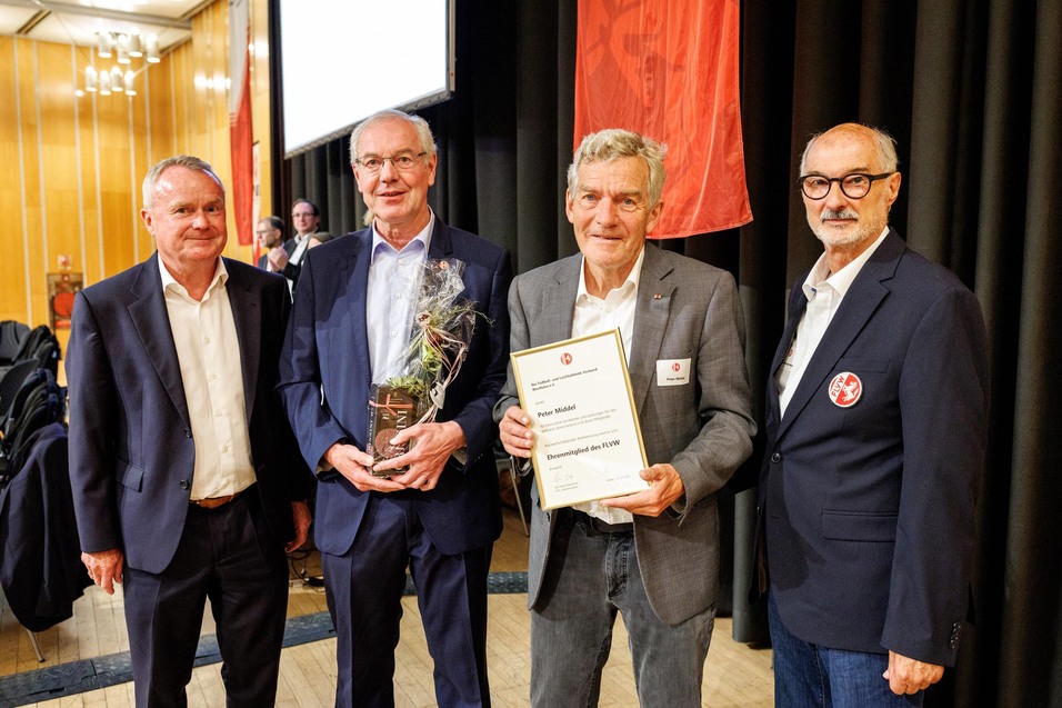 Vier M&auml;nner stehen nebeneinander und blicken in die Kamera. Peter Middel h&auml;lt eine Ehrenurkunde in der Hand, ein weiterer Mann &uuml;berreicht ihm ein Pr&auml;sent. Alle tragen Anzug und l&auml;cheln.