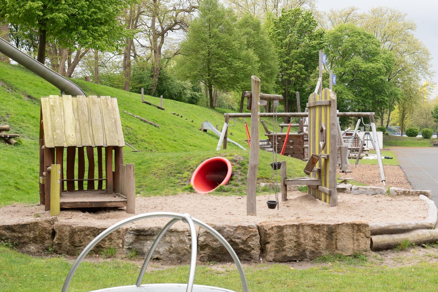 Spielplatz mit Rutsche, Schaukeln und Sandbereich, umgeben von gr&uuml;nen B&auml;umen und Rasenfl&auml;chen.