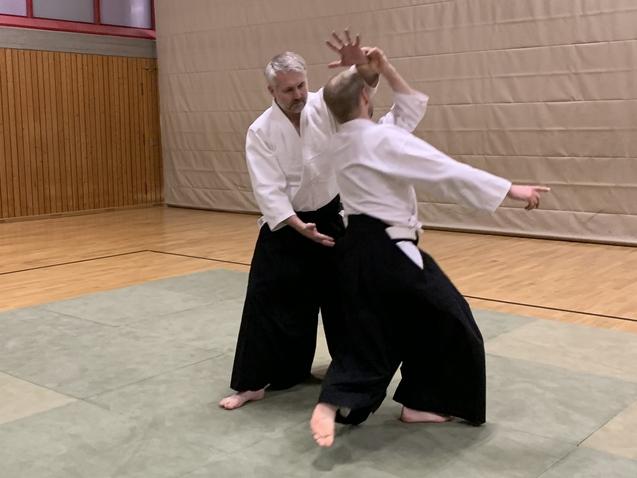 Zwei Personen in traditioneller Aikido-Gewandung demonstrieren eine Technik auf einer Tatami-Matte in einem Sportraum.