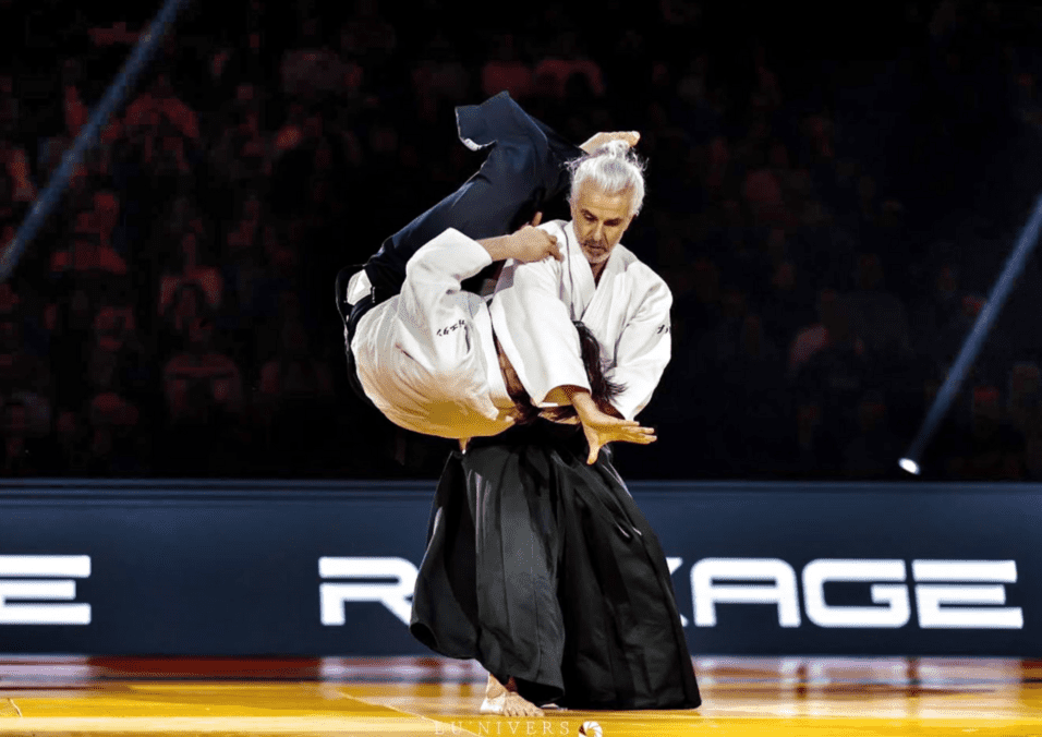 Zwei Kämpfer in traditioneller Kampfkleidung führen einen Wurf während eines Judo-Matches in einer beleuchteten Arena aus.
