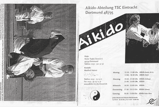 Zwei Kampfkünstler in weißen Anzügen führen eine Aikido-Technik auf einer Matte aus.