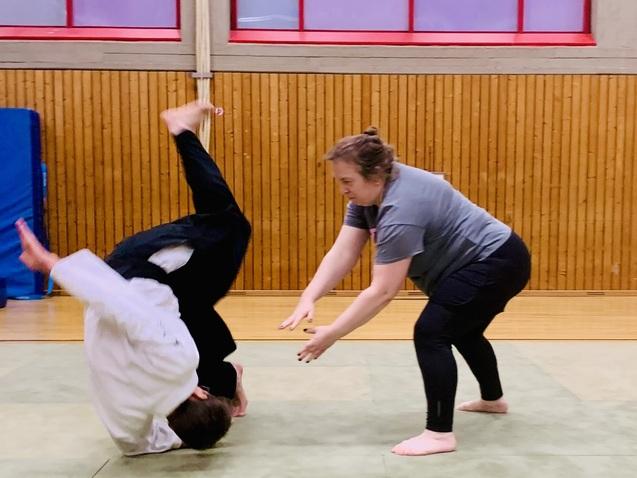 Zwei Personen in Trainingskleidung &uuml;ben eine akrobatische Bewegung auf einer Matte in einer Sporthalle.