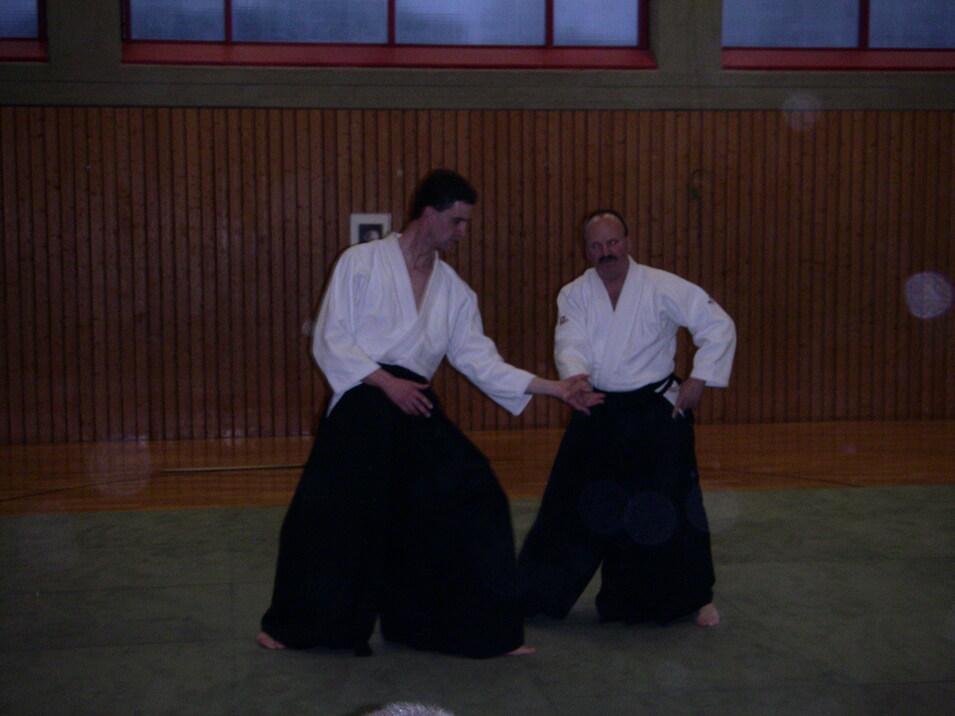 Zwei Männer in traditioneller Bujutsu-Kleidung zeigen eine Technik auf einer Tatami-Matte in einem Trainingsraum.