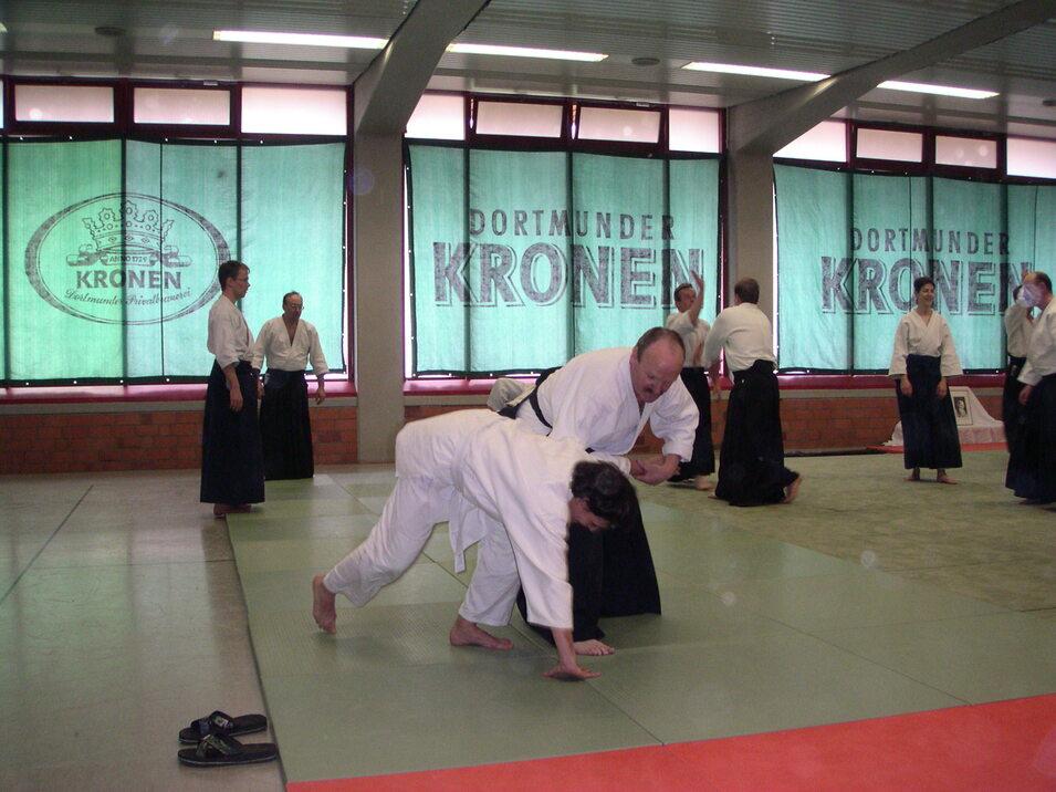 Aikido-Training in einer Halle: Lehrer zeigt einem Schüler eine Technik auf einer Tatami-Matte.