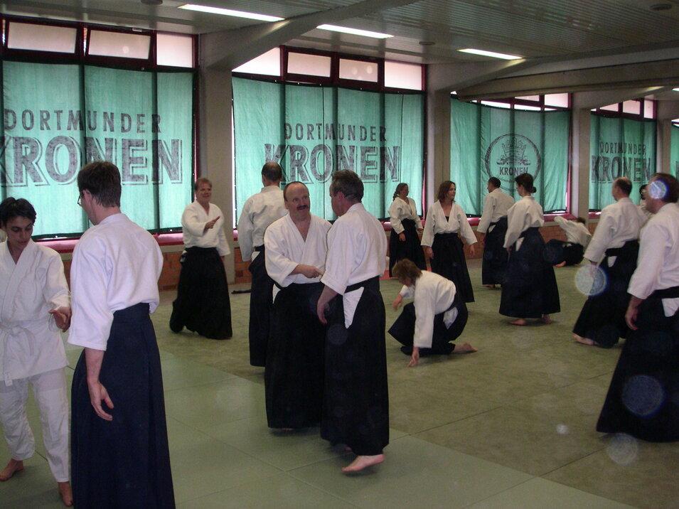 Trainingsszene mit mehreren Personen in weißen Kimonos und schwarzen Hosen, die Aikido in einem großen Raum praktizieren.