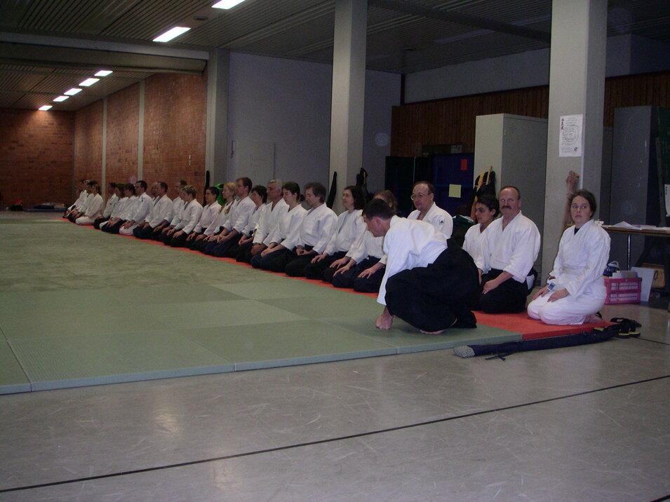 Gruppe von Menschen in weißen Kimonos sitzt auf einer Matte in einer Trainingshalle für Kampfkunst.