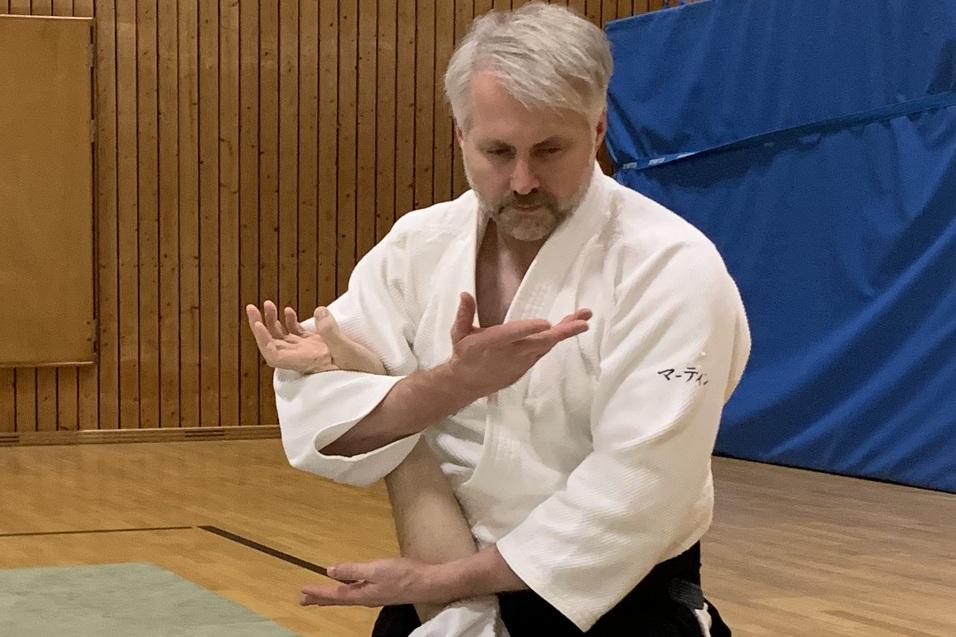 Zwei Personen in weißen Judo-Anzügen auf einer Tatami; einer demonstriert eine Technik auf den anderen.