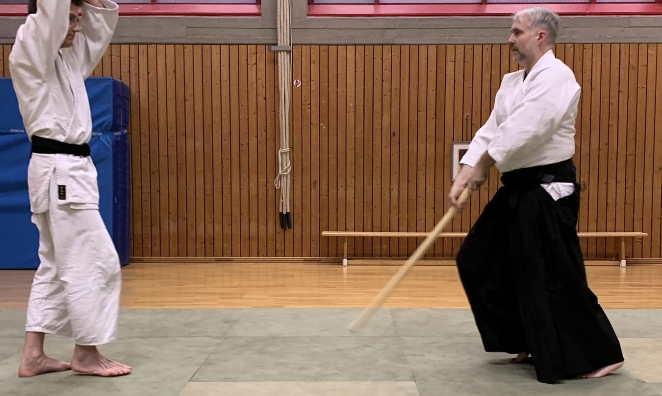 Zwei Kampfsportler in Wei&szlig; und Schwarz &uuml;ben mit einem Holzstock auf einer Tatami-Matte in einem Trainingsraum.