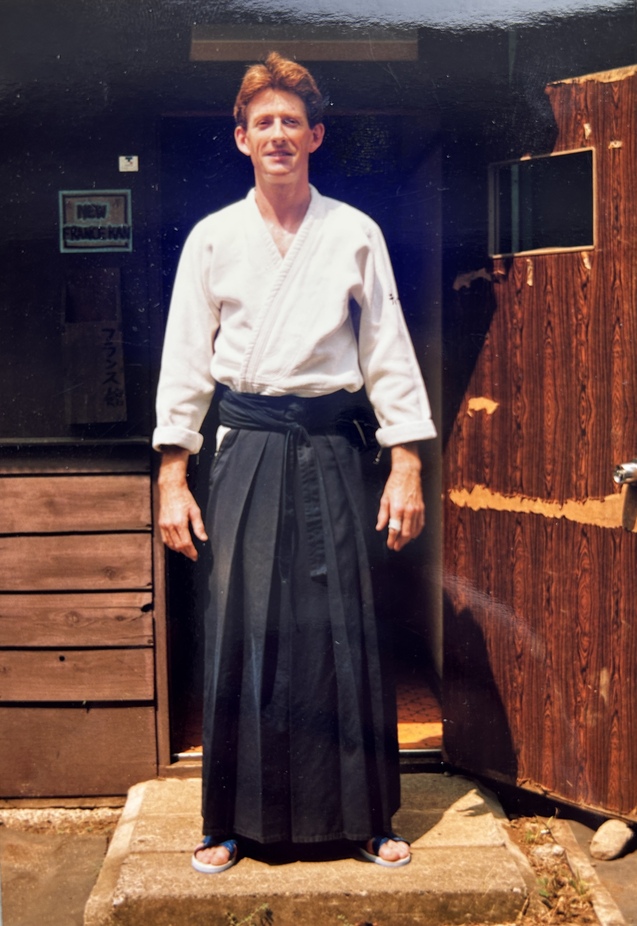 Mann in Hakama und Gi steht vor einem Holzgeb&auml;ude mit einer offenen T&uuml;r und gr&uuml;nem Schild im Hintergrund.