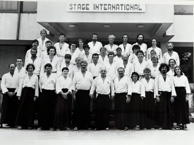 Gruppenfoto von 40 Personen in wei&szlig;en Kimonos und schwarzen Hosen vor einem Geb&auml;ude mit der Aufschrift &bdquo;STAGE INTERNATIONAL&ldquo;.