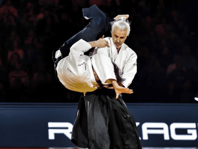 Zwei K&auml;mpfer in traditioneller Kampfkleidung f&uuml;hren einen Wurf w&auml;hrend eines Judo-Matches in einer beleuchteten Arena aus.