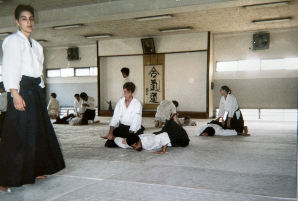 Trainingsszene im Aikido, mehrere Personen auf Tatami, Lehrer erkl&auml;rt Techniken, kalligrafisches Wandbild im Hintergrund.