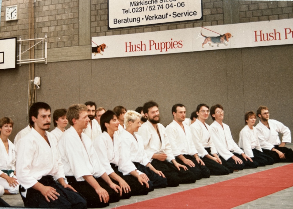 Gruppensitzung von Aikidokas in wei&szlig;en Anz&uuml;gen, auf roten Matten in einem Sportraum.