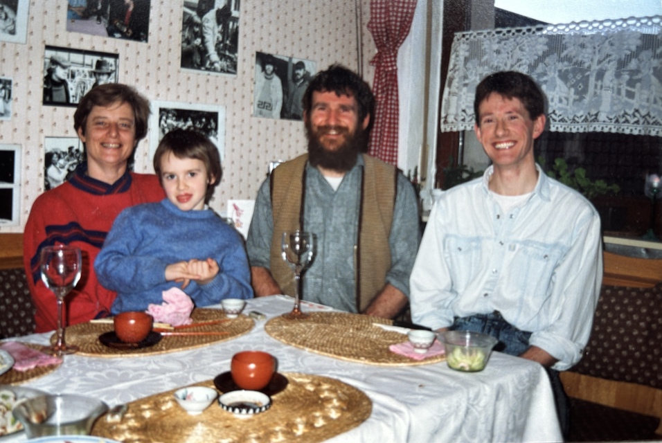Familie sitzt an einem gedeckten Tisch mit Tellern und Gl&auml;sern. Fotos h&auml;ngen an der Wand im Hintergrund.
