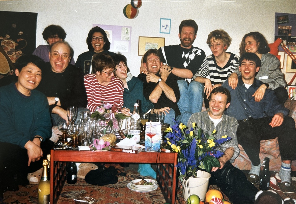 Gruppenkollision von 12 Personen auf einem Sofa, umgeben von Getr&auml;nken und Blumen in einer Hausumgebung.
