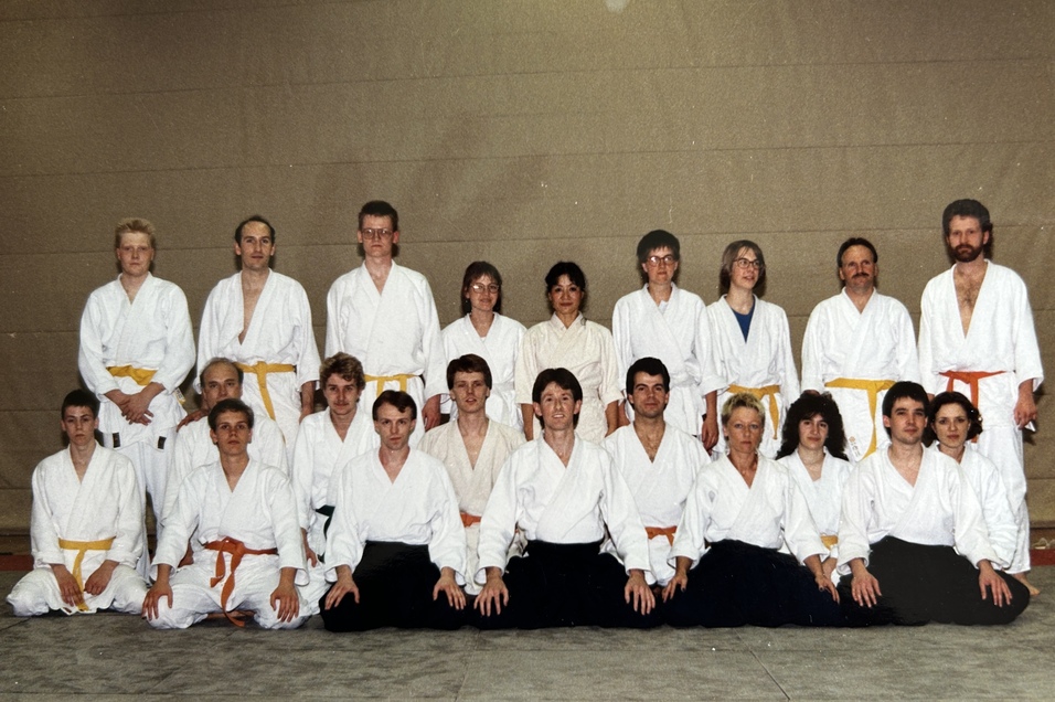 Gruppenshot von 20 Personen in wei&szlig;er und orangefarbener Kampfu attire auf einer Matte in einem Trainingsraum.