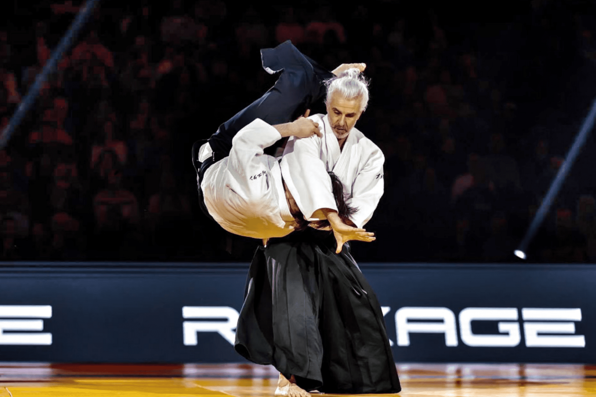 Zwei K&auml;mpfer in traditioneller Kampfkleidung f&uuml;hren einen Wurf w&auml;hrend eines Judo-Matches in einer beleuchteten Arena aus.