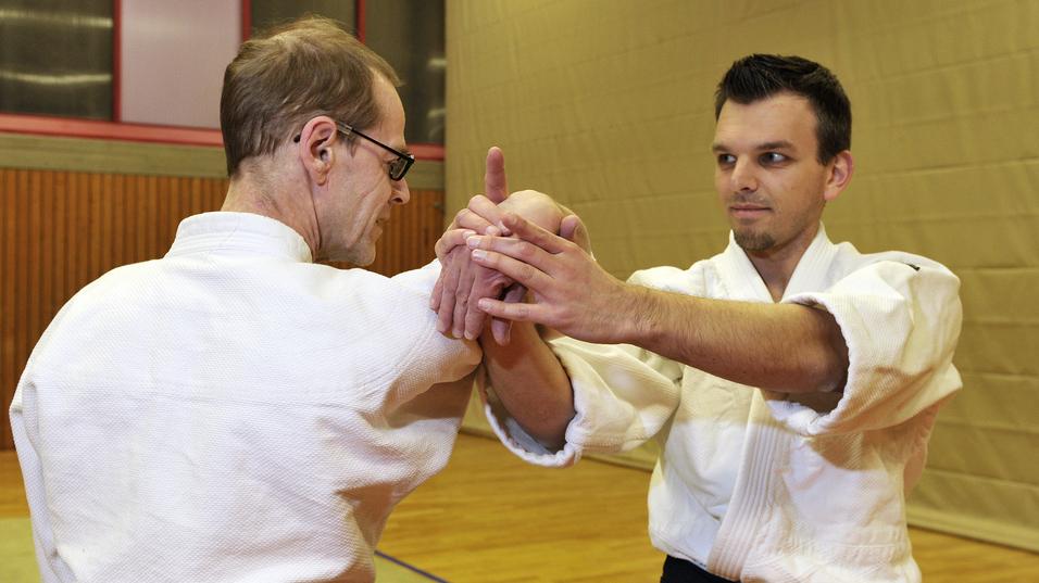 Zwei M&auml;nner im Judogi &uuml;ben eine Technik im Aikido in einer Sporthalle mit Holzboden.