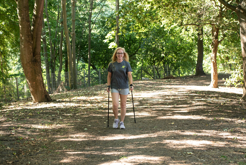 Frau mit Gehst&ouml;cken geht auf einem schmalen Pfad durch einen lichtdurchfluteten Wald.