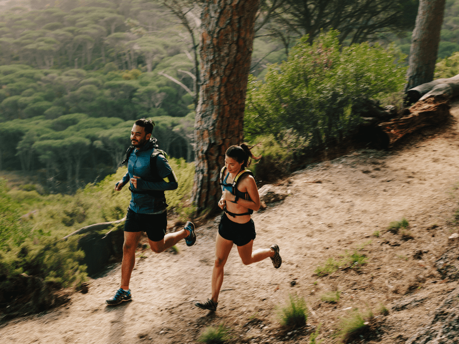 Zwei L&auml;ufer, ein Mann und eine Frau, joggen einen schmalen Pfad durch einen bewaldeten Hang.