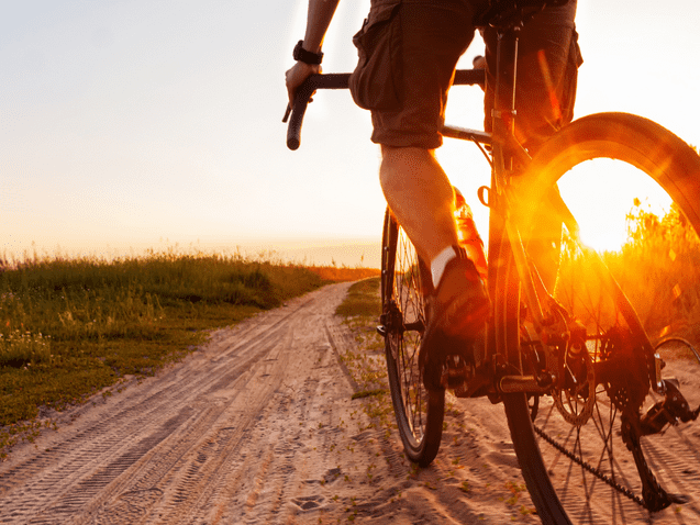 Radfahrer auf einem sandigen Weg bei Sonnenuntergang, umgeben von grüner Vegetation.