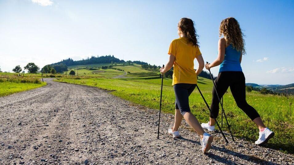 Zwei Frauen wandern mit Nordic-Walking-St&ouml;cken auf einem Kiesweg in einer gr&uuml;nen Landschaft bei Sonnenschein.