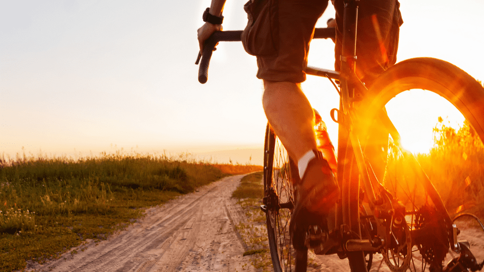 Radfahrer auf einem sandigen Weg bei Sonnenuntergang, umgeben von gr&uuml;ner Vegetation.