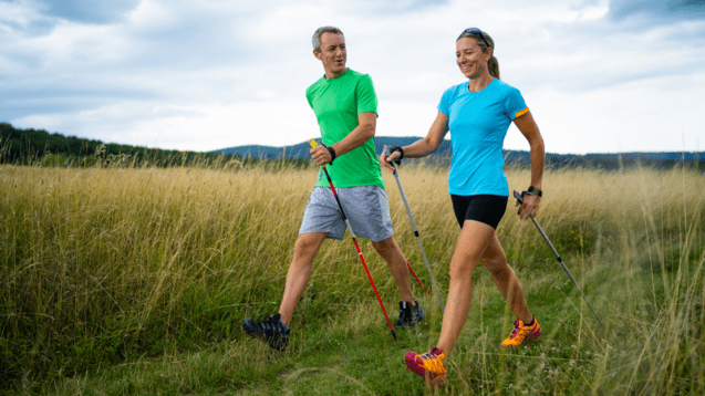 Zweiköpfiges Paar beim Nordic Walking auf einem Feldweg, umgeben von hohem Gras und bewölktem Himmel.