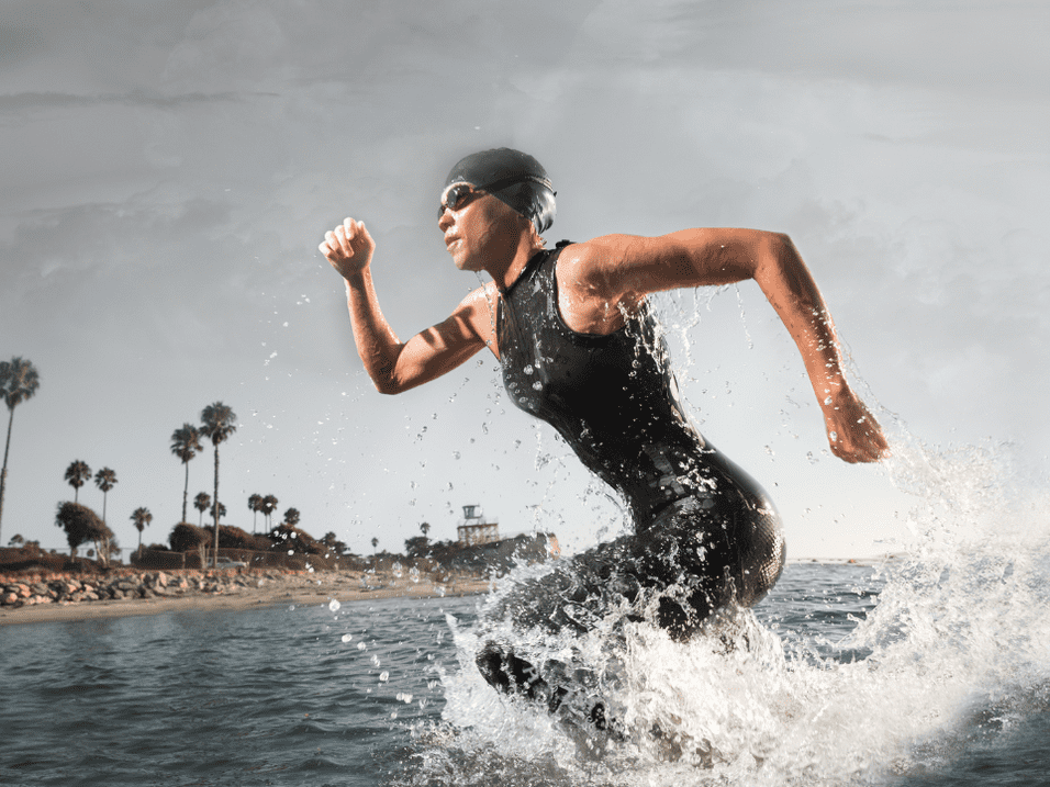 Sportler in schwarzem Schwimmanzug sprintet &uuml;ber das Wasser, diesmal mit Palmen und K&uuml;ste im Hintergrund.