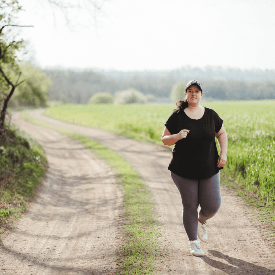 Eine joggende Frau in Sportkleidung auf einem Schotterweg umgeben von grünen Wiesen und Bäumen im Hintergrund.