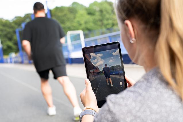 Eine Person filmt mit einem Tablet einen joggenden Mann auf einer Laufbahn im Freien.