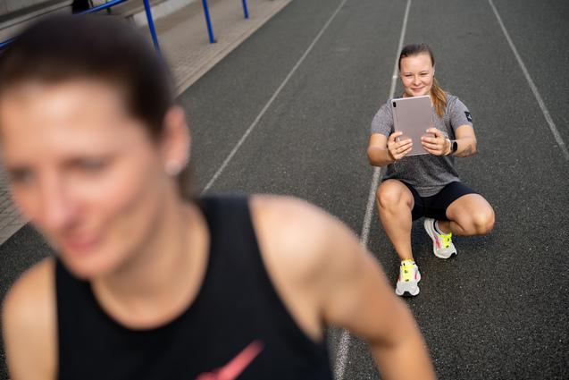 Sportlerin auf einer Laufbahn sitzt und h&auml;lt ein Tablet, w&auml;hrend eine weitere Person im Vordergrund unscharf joggt.