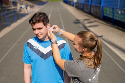 Junge erh&auml;lt ein Ohrstecker-System, w&auml;hrend er auf einer Sportbahn steht. Trainerin hilft ihm dabei.
