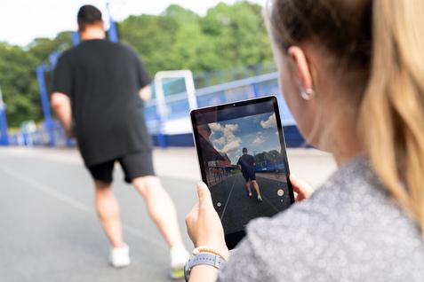 Eine Person filmt mit einem Tablet einen joggenden Mann auf einer Laufbahn im Freien.