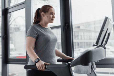 Frau auf einem Crosstrainer in einem Fitnessstudio, blickt auf das Display vor gro&szlig;en Fenstern.