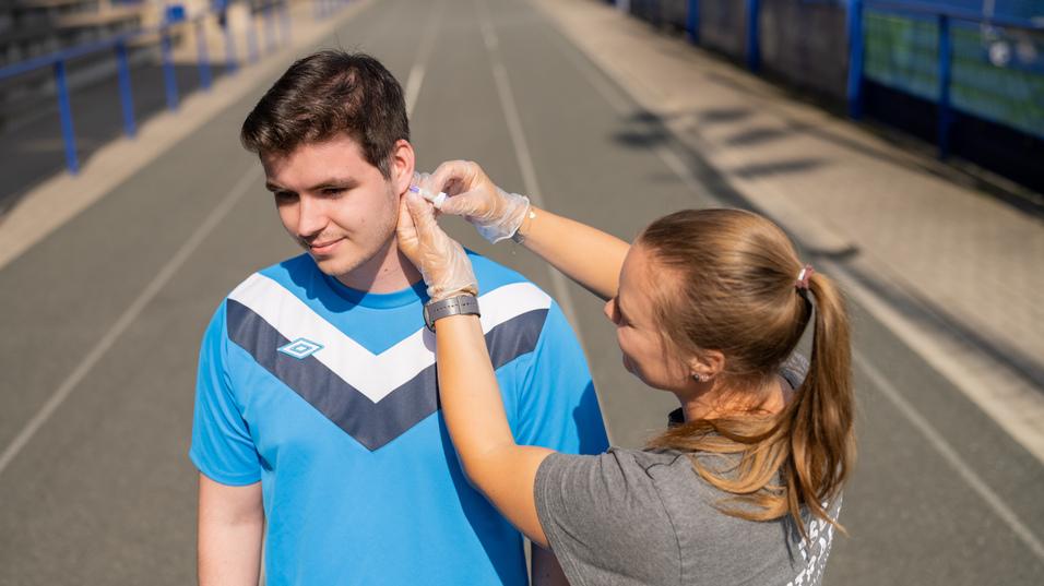 Junge erhält ein Ohrstecker-System, während er auf einer Sportbahn steht. Trainerin hilft ihm dabei.