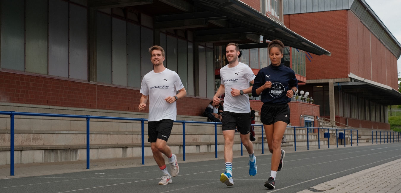 Drei L&auml;ufer in Sportkleidung joggen auf einer Leichtathletikanlage entlang eines blauen Gel&auml;nders.