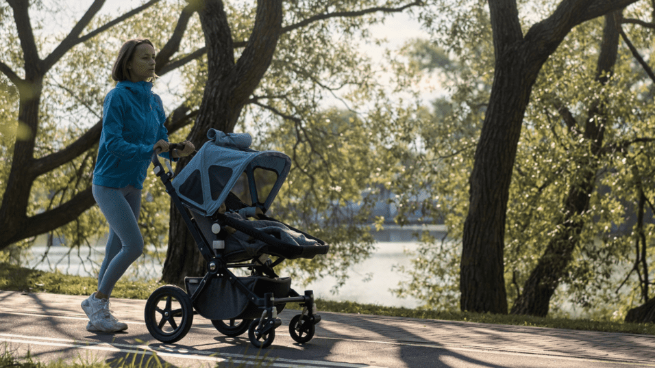 Eine joggende Frau schiebt einen Kinderwagen auf einem Weg entlang eines gr&uuml;nen Ufers. Sonnenschein durch B&auml;ume.