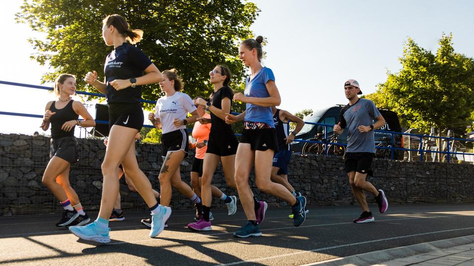 Gruppe von Joggern in Sportkleidung, die auf einem Gehweg bei sonnigem Wetter entlanglaufen.
