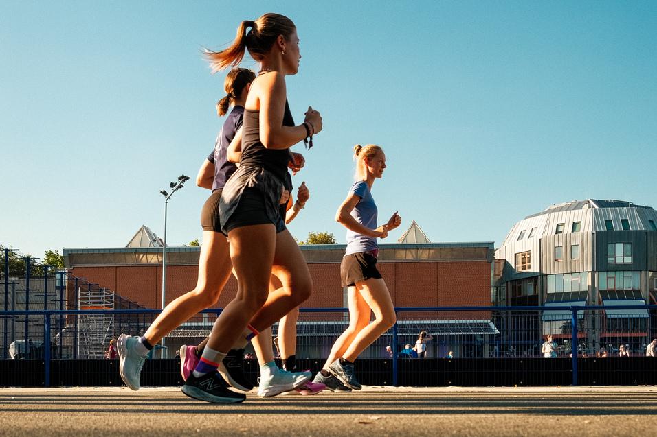 Vier Frauen joggen auf einem Asphaltweg unter klarem Himmel, im Hintergrund moderne Gebäude sichtbar.
