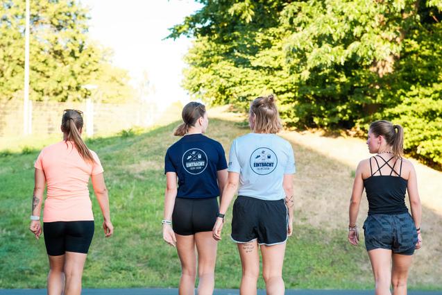 Vier Frauen in Sportkleidung gehen Rücken an Rücken auf einem Weg zwischen Bäumen.