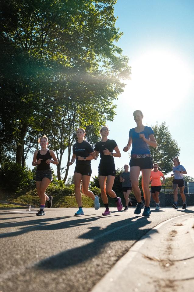 Gruppe von Frauen joggt auf einem Gehweg unter strahlendem Sonnenschein, umgeben von B&auml;umen.