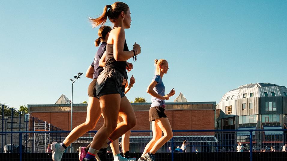 Vier Frauen joggen auf einem Asphaltweg unter klarem Himmel, im Hintergrund moderne Gebäude sichtbar.