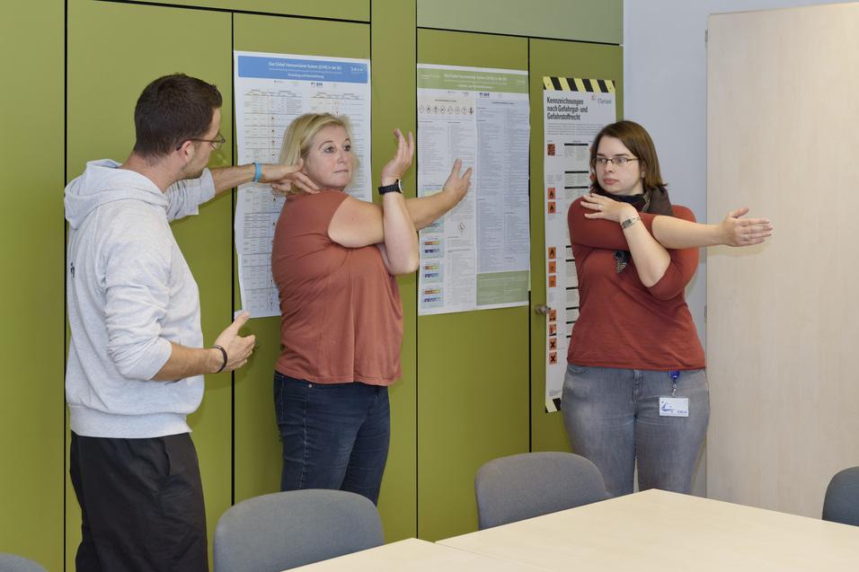 Drei Personen dehnen ihre Arme in einem Raum mit Informationspostern an den Wänden.