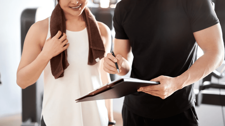 Zwei Personen im Fitnessstudio, eine Frau mit Handtuch, bespricht mit einem Trainer auf einem Clipboard seine Anweisungen.