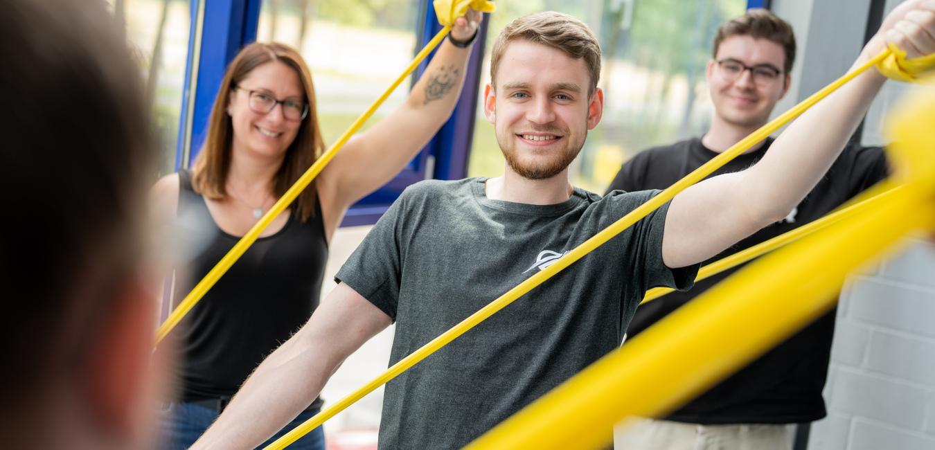 Drei Personen trainieren mit gelben Widerstandsbändern in einem hellen Raum mit großen Fenstern.