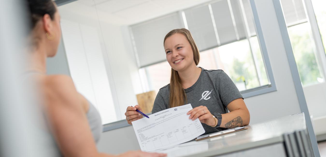 Zwei Frauen sitzen an einem Tisch; eine lächelt und zeigt auf ein Dokument, während die andere aufmerksam zuhört.
