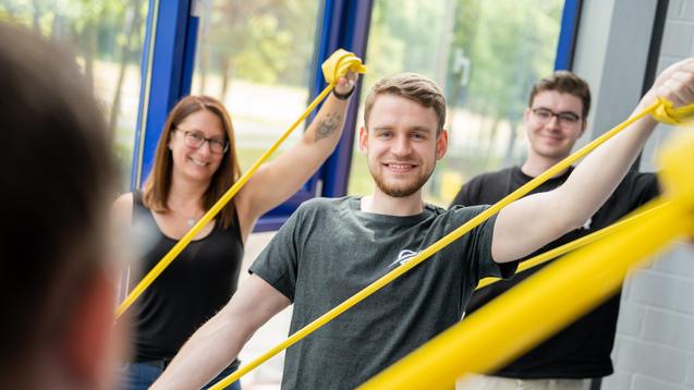 Drei Personen trainieren mit gelben Widerstandsbändern in einem hellen Raum mit großen Fenstern.